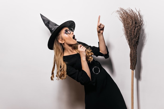 Lockiges Mädchen im Halloween-Kostüm, das oben schaut. Charmante Hexe im schwarzen Hut, der mit altem Besen aufwirft.