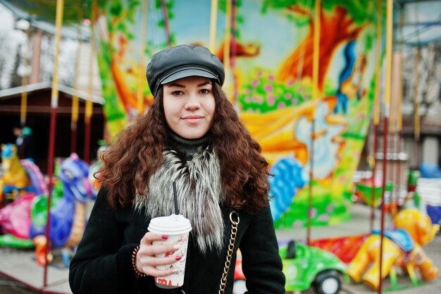 Lockiges mexikanisches Mädchen in Lederkappe und Plastiktasse Kaffee zur Hand gegen Karussell