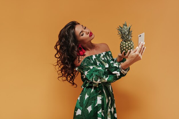 Lockiges modernes Mädchen mit dunklen langen Haaren und großen Lippen in roten Ohrringen und bedrucktem Sommerkleid, das Selfie macht und Ananas hält holding