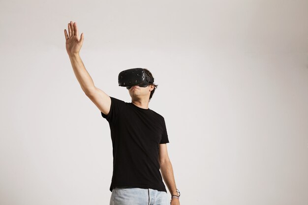 Low-Angle-Porträt eines jungen kaukasischen Modells in Jeans und unbeschriftetem schwarzem T-Shirt, das seine Hand ausstreckt, während in VR-Brille auf weißer Wand spielt.