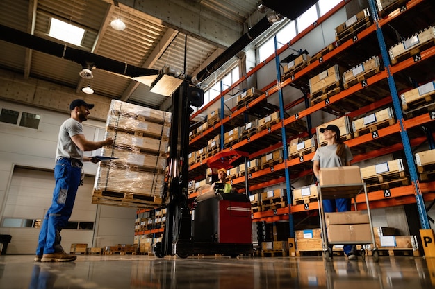 Low Angle View von glücklichen Lagerarbeitern, die kommunizieren, während sie mit dem Versand im industriellen Lagerabteil arbeiten