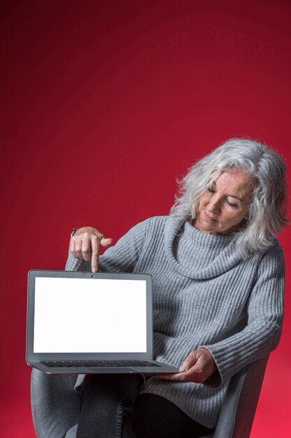 Ältere Frau, die auf dem Lehnsessel zeigt ihren Finger auf einem offenen Laptop sitzt