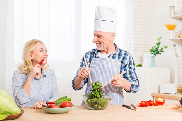 Ältere Frau, die den roten Paprika beißt, der den Salat in der Küche zubereitend betrachtet