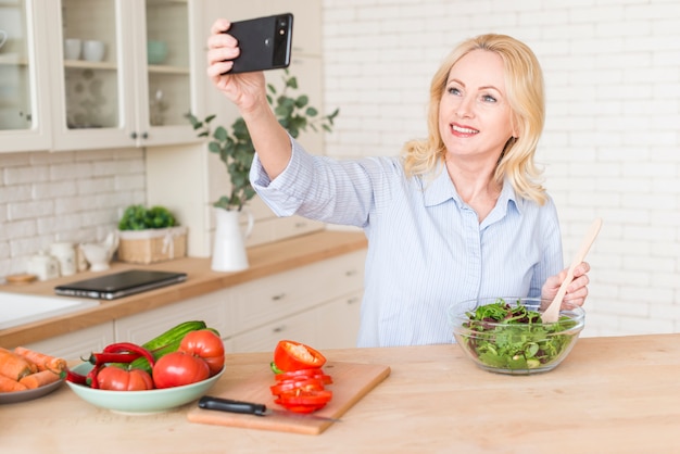 Ältere Frau, die den Salat nimmt selfie am Handy zubereitet