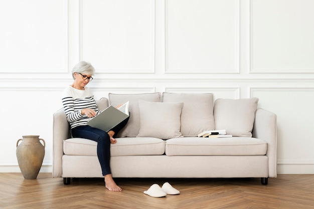 Ältere Frau, die ein Buch auf dem Sofa in einem skandinavischen Dekor-Wohnzimmer liest