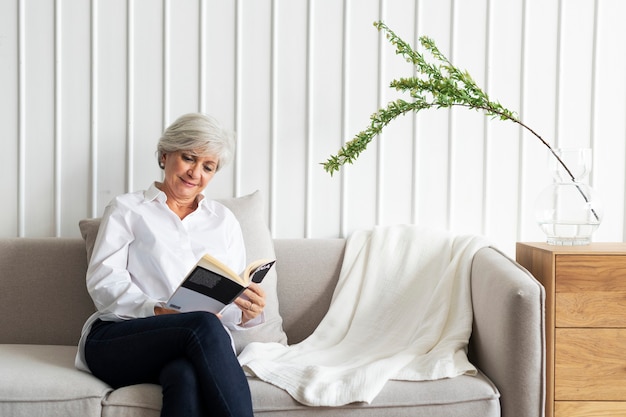 Ältere Frau, die ein Buch auf dem Sofa in einem skandinavischen Dekor-Wohnzimmer liest