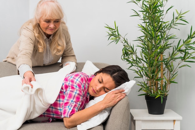 Ältere Frau, die eine Decke auf ihre erschöpfte Tochter schlafend auf das Sofa setzt