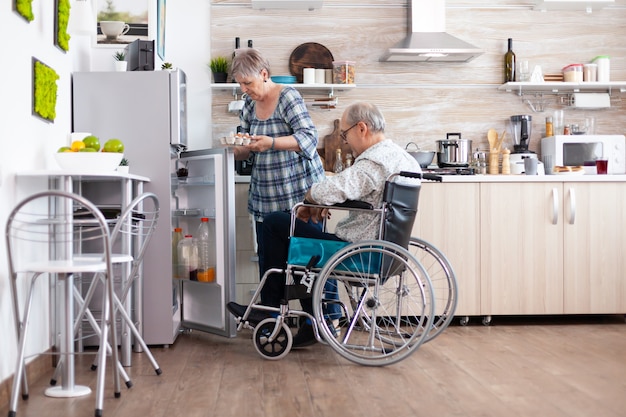 Ältere Frau, die Frühstück für behinderten Ehemann zubereitet, der Eierkarton aus dem Kühlschrank nimmt und mit einem Mann mit Gehbehinderung lebt. Behinderter älterer Mann im Rollstuhl, der seiner Frau in der Küche hilft
