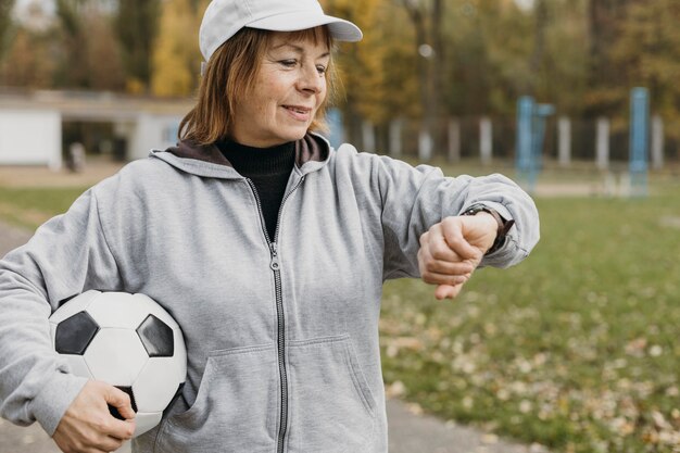 Ältere Frau, die Fußball hält und ihre Uhr im Freien betrachtet