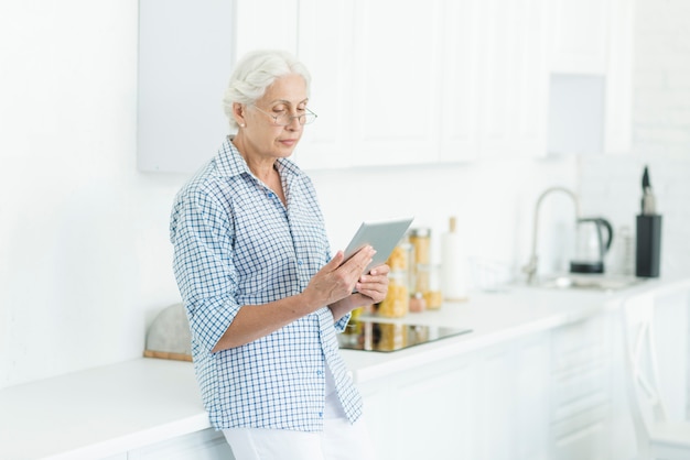 Ältere Frau, die in der Küche betrachtet digitale Tablette steht