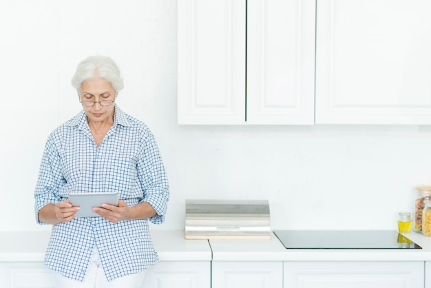 Ältere Frau, die in der Küche unter Verwendung der digitalen Tablette steht