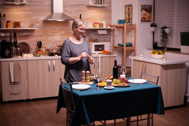 Ältere Frau, die Kerzen in der Küche für ein romantisches Abendessen mit Ehemann anzündet. Ältere Frau, die ihren Ehemann auf ein romantisches Abendessen wartet. Reife Frau, die festliches Essen für die Jubiläumsfeier zubereitet.