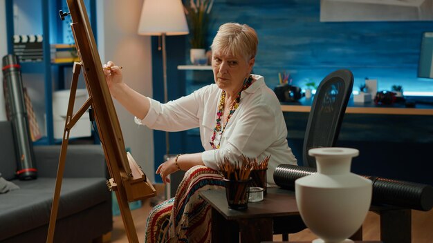 Ältere Frau, die Kunstwerkskizze mit künstlerischen Fähigkeiten und Bleistiften auf weißer Leinwand mit hölzerner Staffelei zeichnet. Verwenden Sie Ausrüstung und Vase als Inspiration, um professionelle Meisterwerke zu zeichnen. Handschuss.