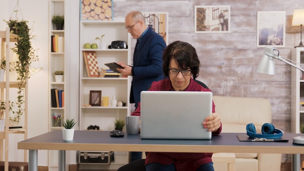 Ältere Frau, die Laptop öffnet und seine Brille auf ihr Gesicht setzt. Alter Mann mit Brille mit Tablet im Brackground.