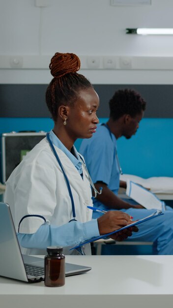 Ältere Frau, die medizinischen Rat von einer Krankenschwester erhält, während sie im Schrank der Einrichtung auf dem Bett sitzt. Junger Arzt spricht mit älteren Patienten am Schreibtisch, während er Informationen zur Untersuchung schreibt