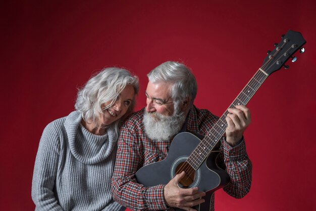 Ältere Frau, die nahe ihrem Ehemann spielt die Gitarre gegen roten Hintergrund sitzt