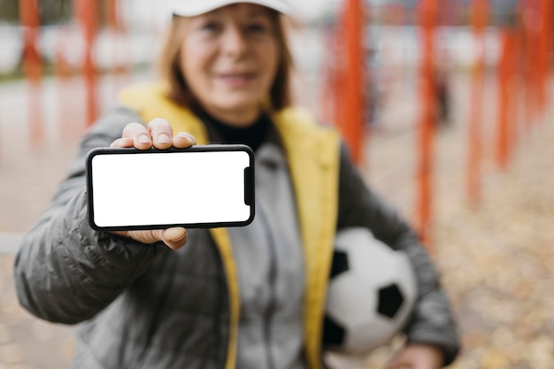 Ältere Frau, die Smartphone und Fußball beim Training im Freien hält