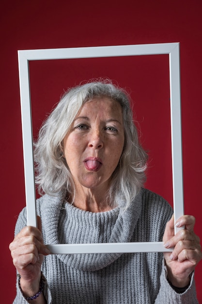 Ältere Frau, die weiße Rahmengrenze heraus haftet ihre Zunge gegen roten Hintergrund hält