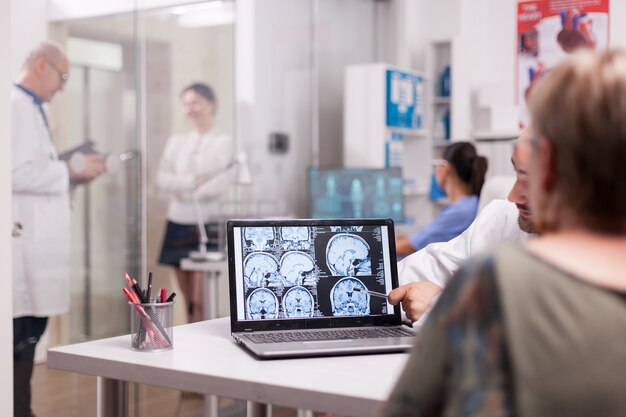 Ältere Frau im Krankenhausbüro, die sich den CT-Scan des Gehirns ansieht, während sie mit dem Arzt über die Diagnose spricht. Kranke junge Frau und älterer Mediziner mit grauem Haar im Klinikkorridor.