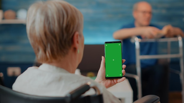 Ältere Frau im Rollstuhl, die zu Hause ein Smartphone mit grünem Bildschirm hält. Rentner mit chronischer Behinderung mit isoliertem Kopierraum mit leerem Mock-up und Chroma-Key-Hintergrund auf dem Display.