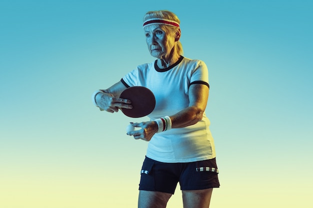 Ältere Frau im Sportbekleidungstraining im Tischtennis auf Gradientenhintergrund, Neonlicht. Weibliches Model in guter Form bleibt aktiv. Konzept von Sport, Aktivität, Bewegung, Wohlbefinden, Selbstvertrauen. Copyspace.