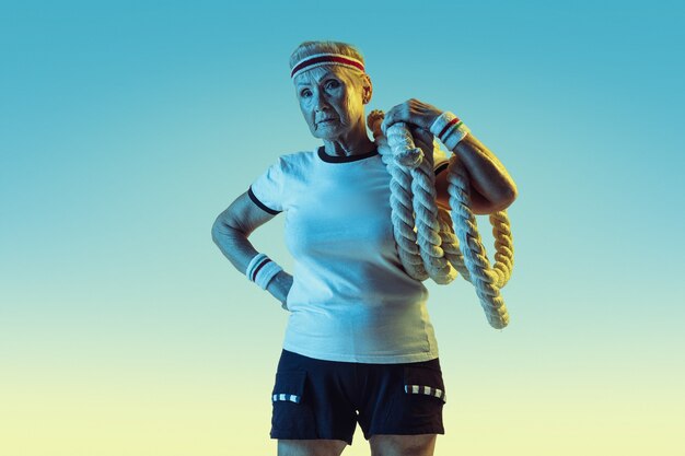 Ältere Frau im Sportbekleidungstraining mit Seilen auf Gradientenhintergrund, Neonlicht.