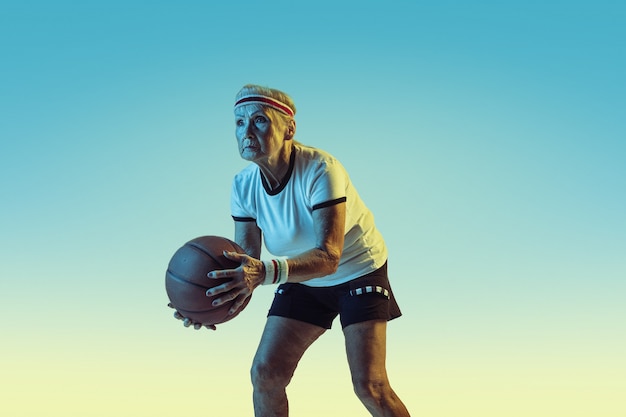 Ältere Frau in der Sportbekleidung, die Basketball auf Gradientenhintergrund, Neonlicht spielt.
