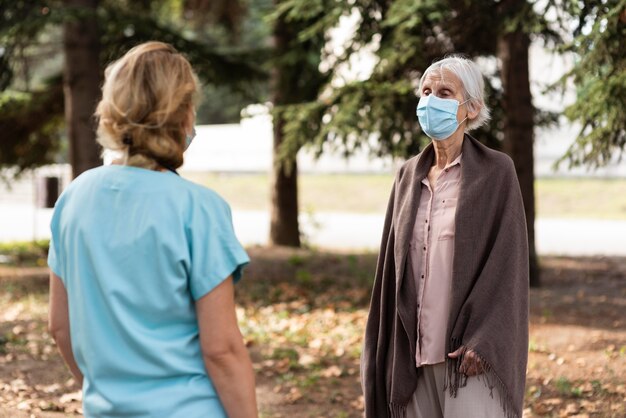 Ältere Frau mit medizinischer Maske im Gespräch mit weiblicher Krankenschwester im Freien