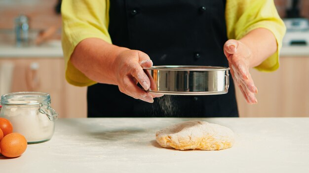 Ältere Frau mit schwarzer Schürze, die Mehl auf Teig mit metallischem Sieb durchsiebt Glücklicher älterer Koch mit Bone, der Rohzutaten zum Backen von traditionellem Brotbesprengen, Sieben in der Küche zubereitet.