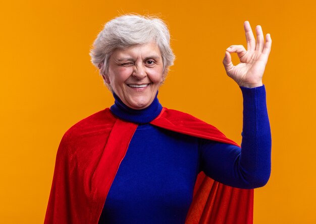Ältere Frau Superheld mit rotem Umhang, der die Kamera lächelt und zwinkert und ein ok-Zeichen auf orangefarbenem Hintergrund zeigt