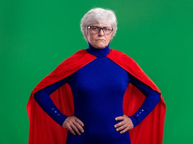 Ältere Frau Superheldin mit rotem Umhang und Brille, die Kamera mit ernstem Gesicht mit Armen an der Hüfte über grünem Hintergrund anschaut