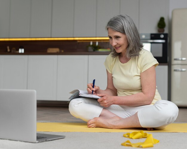 Ältere Frau zu Hause, die Fitnessunterricht auf dem Laptop studiert und Notizen macht