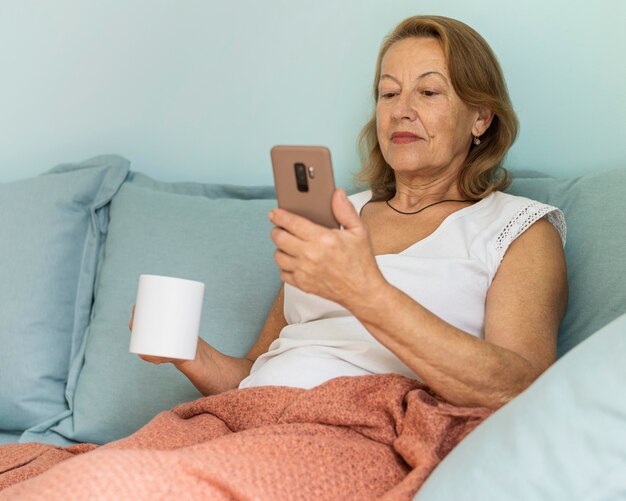 Ältere Frau zu Hause während der Pandemie, die eine Tasse Kaffee genießt und Smartphone verwendet