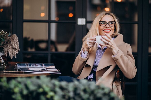 Ältere Geschäftsfrau in einem Mantel, der außerhalb des Cafés sitzt und Zeitschrift liest