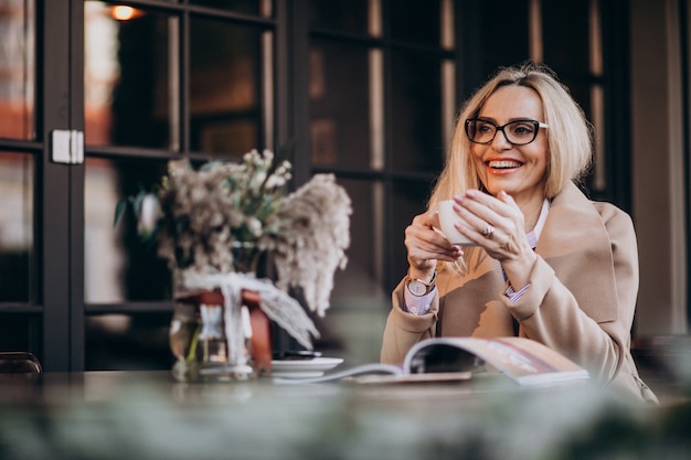 Ältere Geschäftsfrau in einem Mantel, der außerhalb des Cafés sitzt und Zeitschrift liest