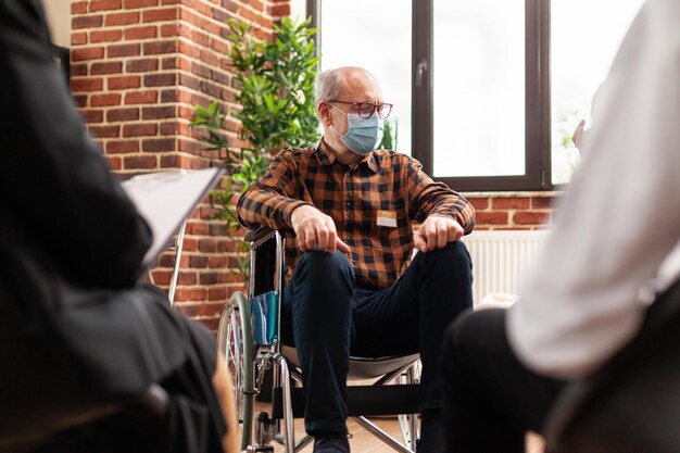 Ältere Person mit Behinderung, die an einer Gruppentherapiesitzung teilnimmt und eine Gesichtsmaske trägt. Mann sitzt im Rollstuhl bei einem Meeting-Programm, um Beratung und Ratschläge gegen Alkoholsucht zu erhalten.