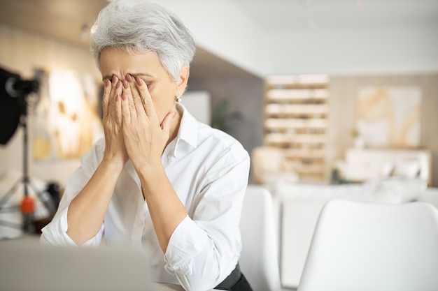 Ältere traurige Frau mit grauem Haar, das am Laptop arbeitet, Augen reibt oder Tränen versteckt, voller unruhiger Gedanken