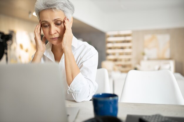 Ältere traurige Frau mit grauem Haar, das am Laptop arbeitet, Augen reibt oder Tränen versteckt, voller unruhiger Gedanken