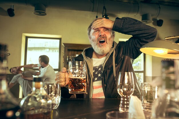 Älterer bärtiger Mann, der Alkohol in der Kneipe trinkt und ein Sportprogramm im Fernsehen sieht. Bier genießen. Mann mit Bierkrug, der am Tisch sitzt. Fußball- oder Sportfan. Kampf der Fans im Hintergrund