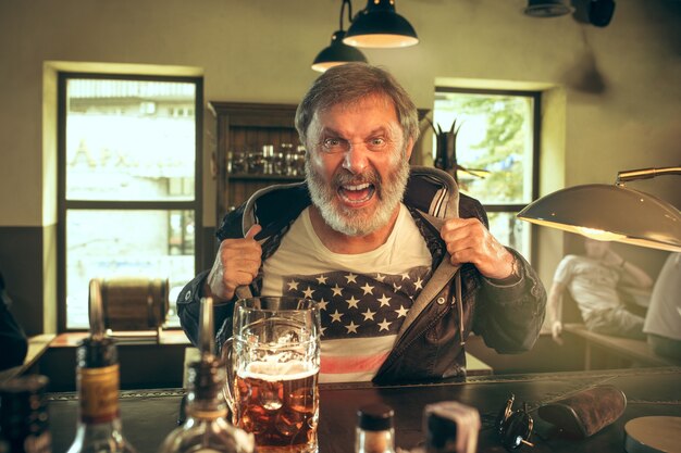 Älterer bärtiger Mann, der Alkohol in der Kneipe trinkt und ein Sportprogramm im Fernsehen sieht. Ich genieße meinen Lieblingsbrot und mein Lieblingsbier. Mann mit Bierkrug, der am Tisch sitzt. Fußball- oder Sportfan. Konzept der menschlichen Emotionen