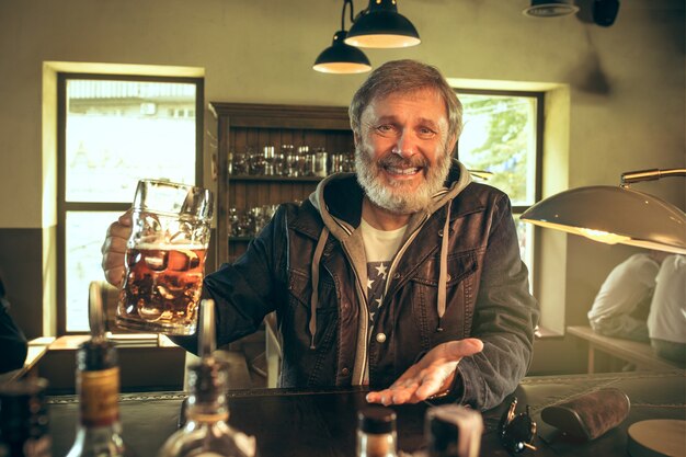 Älterer bärtiger Mann, der Alkohol in der Kneipe trinkt und ein Sportprogramm im Fernsehen sieht. Ich genieße meinen Lieblingsbrot und mein Lieblingsbier. Mann mit Bierkrug, der am Tisch sitzt. Fußball- oder Sportfan. Konzept der menschlichen Emotionen