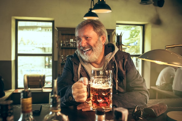 Älterer bärtiger Mann, der Alkohol in der Kneipe trinkt und ein Sportprogramm im Fernsehen sieht. Ich genieße meinen Lieblingsbrot und mein Lieblingsbier. Mann mit Bierkrug, der am Tisch sitzt.