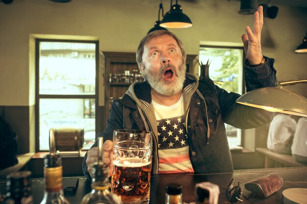 Älterer bärtiger Mann, der Alkohol in der Kneipe trinkt und ein Sportprogramm im Fernsehen sieht.