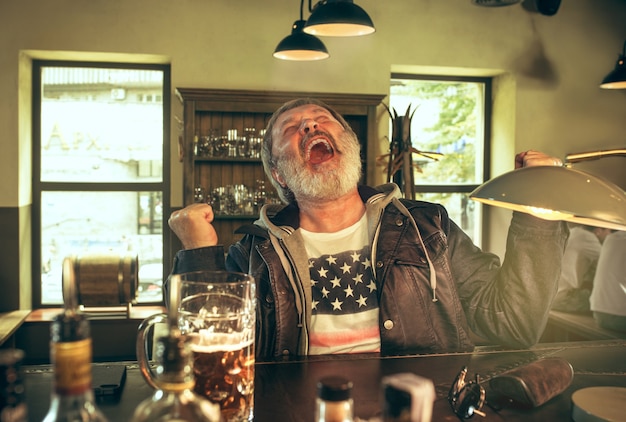 Älterer bärtiger Mann, der Alkohol in der Kneipe trinkt und ein Sportprogramm im Fernsehen sieht.