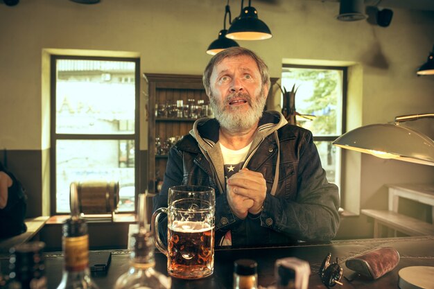 Älterer bettelnder bärtiger Mann, der Alkohol in der Kneipe trinkt und ein Sportprogramm im Fernsehen sieht. Ich genieße meinen Lieblingsbrot und mein Lieblingsbier. Mann mit Bierkrug, der am Tisch sitzt. Fußball- oder Sportfan. Menschliche Gefühle