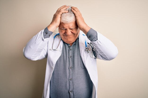 Älterer gutaussehender grauhaariger Arzt mit Mantel und Stethoskop auf weißem Hintergrund, der unter Kopfschmerzen leidet, die verzweifelt und gestresst sind, weil Schmerzen und Migräne Hände auf den Kopf