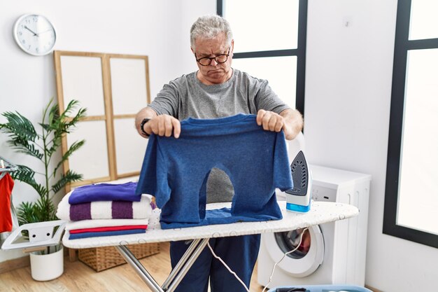 Älterer kaukasischer Mann, der ein verbranntes Eisenhemd in der Waschküche bügelt, entspannt mit ernstem Gesichtsausdruck. einfach und natürlich in die Kamera blicken.