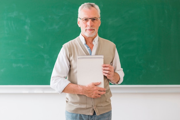 Älterer männlicher Professor, der das Notizbuch steht gegen Tafel hält