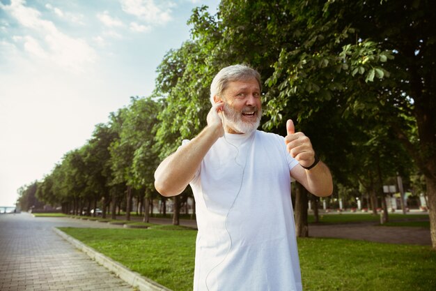 Älterer Mann als Läufer mit Fitness-Tracker an der Stadtstraße. Kaukasisches männliches Modell mit Geräten beim Joggen und Cardio-Training am Sommermorgen. Gesunder Lebensstil, Sport, Aktivitätskonzept.