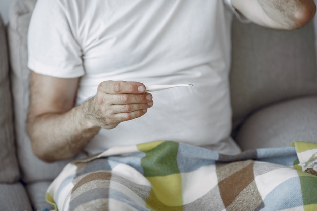 Älterer Mann, der allein auf Sofa sitzt. Kranker Mann mit Plaid bedeckt. Grangfather mit Thermometer.